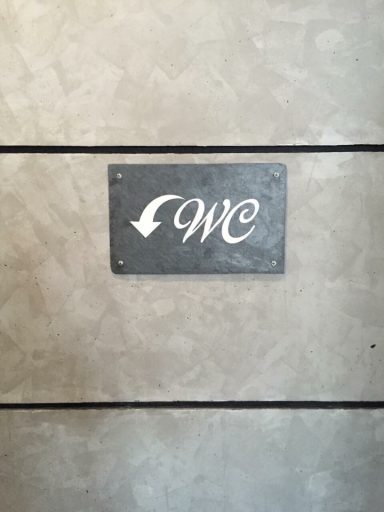 Hinweisschild für die Richtung zum WC, graue Wand mit Pfeil nach unten.