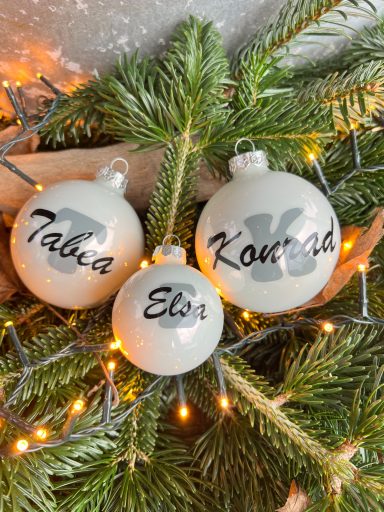 Drei Weihnachtskugeln mit den Namen Tabia, Elia und Kounia auf Tannenzweigen.