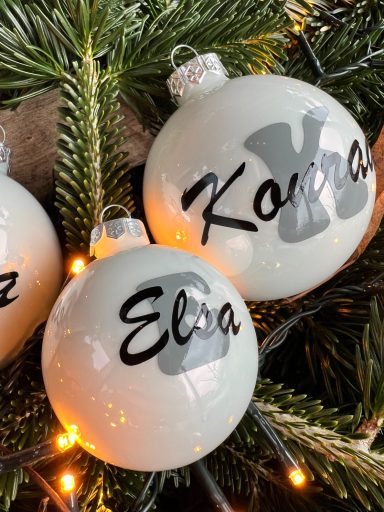 Weihnachtsbaumkugeln mit den Namen "Kaya" und "Elza" auf einem Hintergrund aus Tannenzweigen.